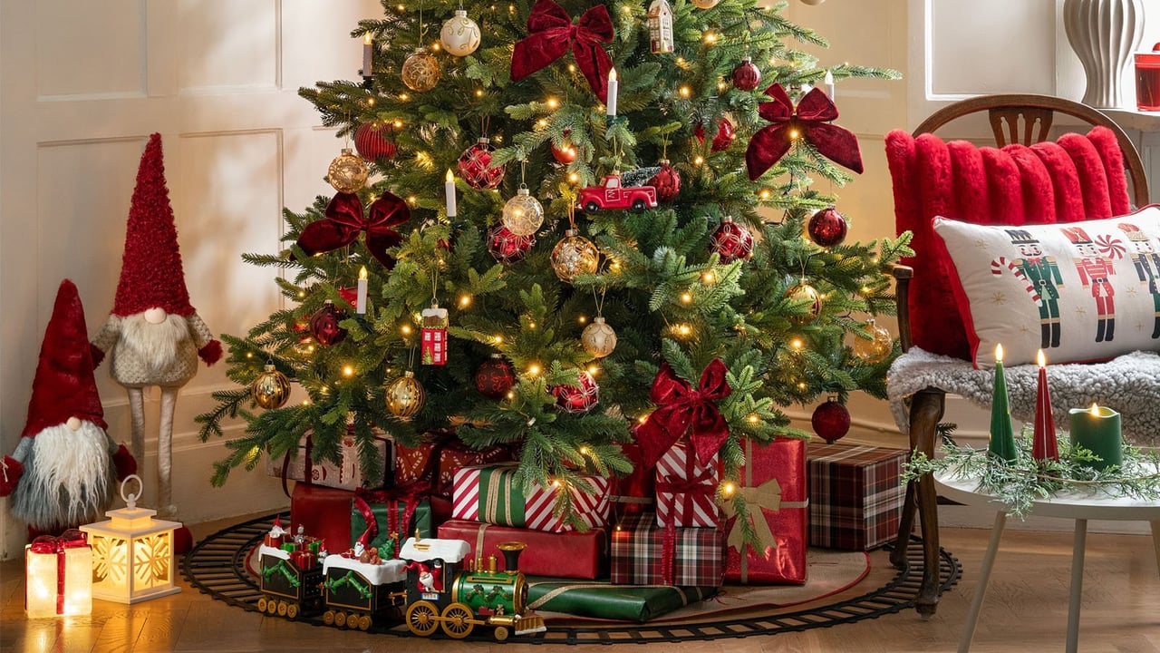 Weihnachtsbaum mit Geschenken, Schmuck und Spielzeugzug in einer gemütlich eingerichteten Ecke mit Wichteln und Kissen.