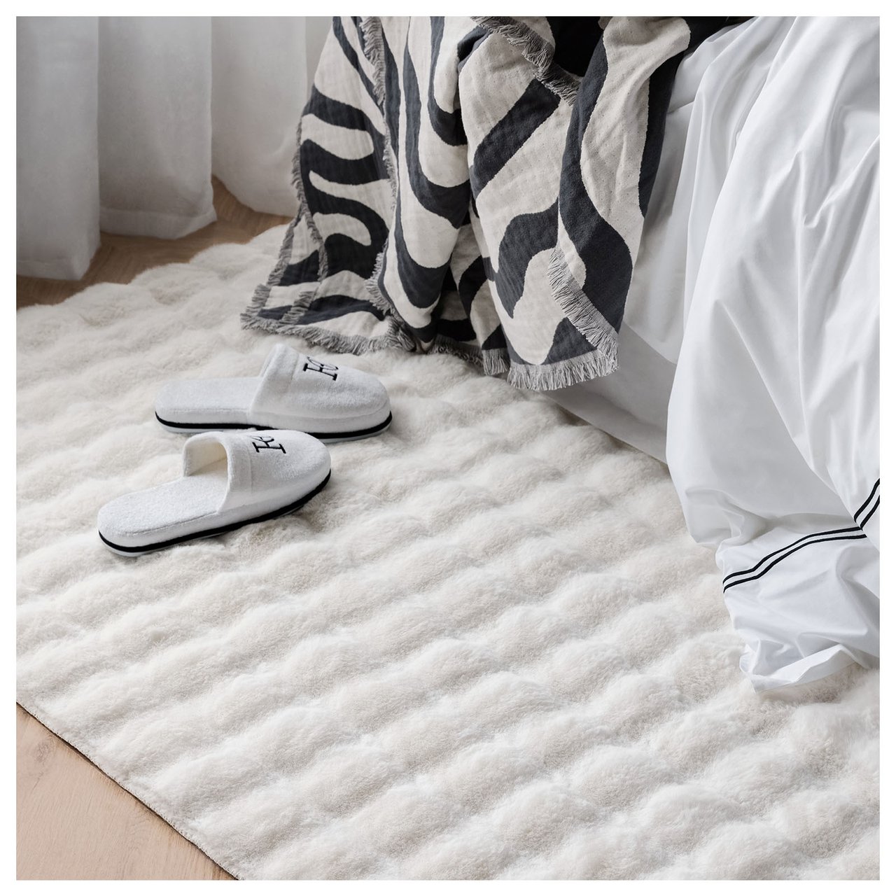 Weißer, flauschiger Teppich unter einem Bett, mit Hausschuhen drauf.