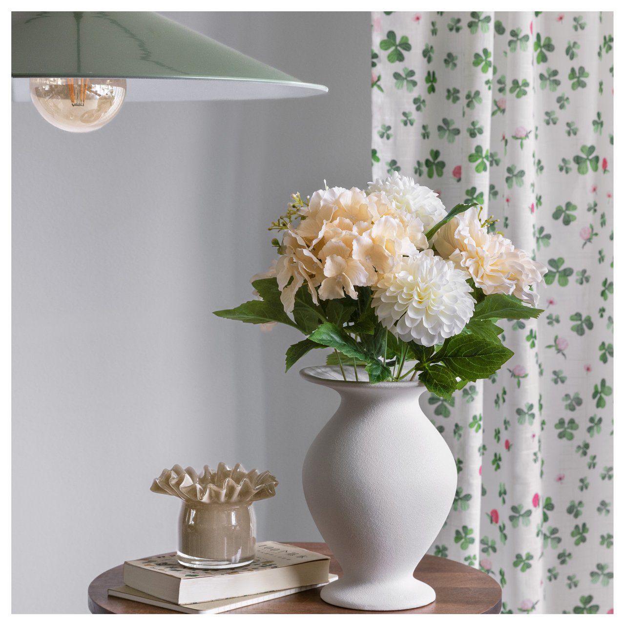 Beistelltisch mit einer Vase, in der Kunstblumen stehen, Buch und Laterne vor einem Vorhang mit grünen Blüten.