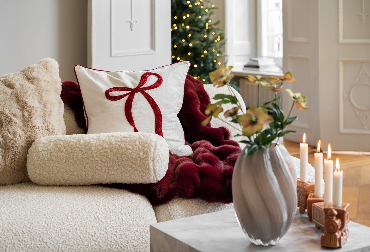 Helles Sofa mit Weihnachtskissen mit roter Schleife, weichen Decken und Kissen. Auf dem Tisch stehen Vase und Kerzen.
