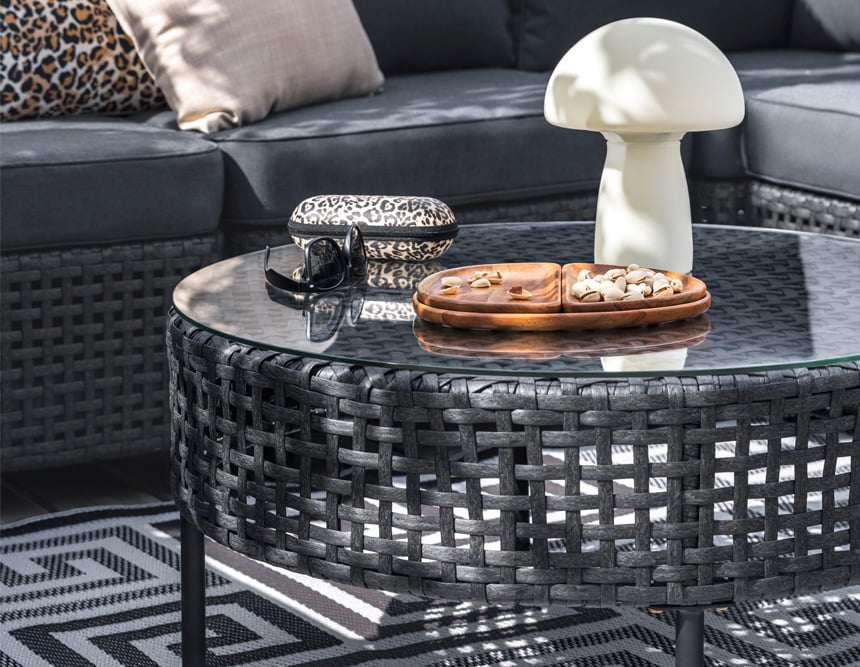 Loungebord i konstrotting med glas, snacks, lampa och solglasögon framför en mörk loungesoffa.