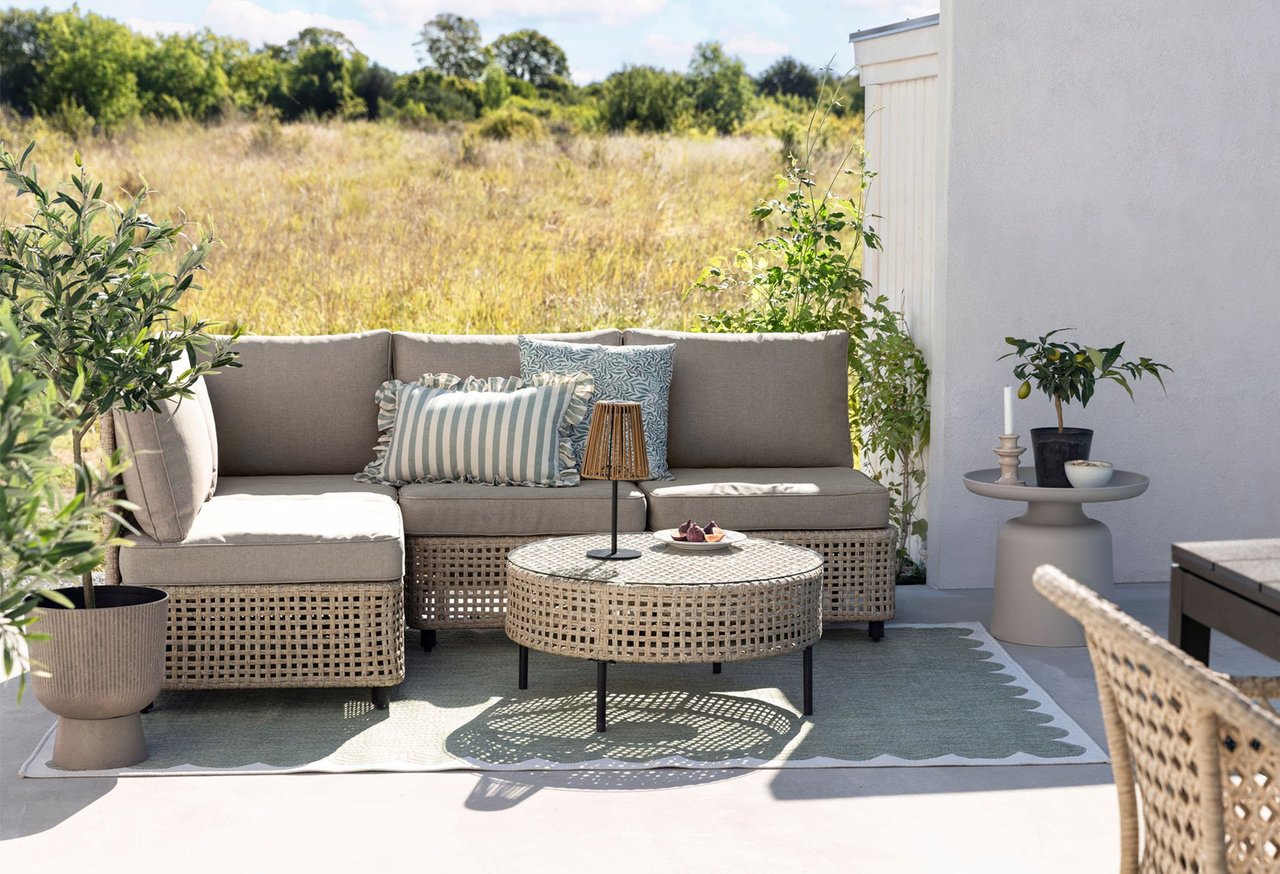 Sonnige Terrasse mit beigem Rattan-Loungesofa, rundem Tisch, Kissen und grünen Pflanzen vor Natur.