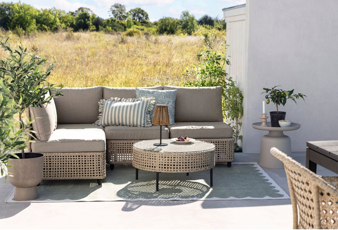 Aurinkoinen terassi, beige rottinkisohva, tyynyjä, pyöreä pöytä ja vihreitä kasveja luonnon edessä.