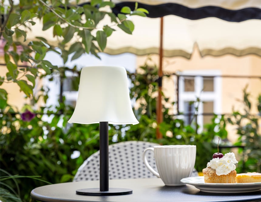 Pöytävalaisin ulkopöydällä kupin ja leivoksen kanssa, vihreiden kasvien ympäröimänä parvekkeella.