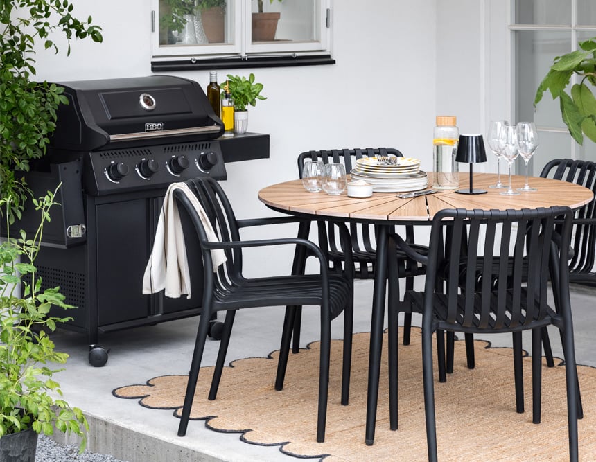 Terassi, jossa musta grilli, pyöreä puupöytä, mustat tuolit, kattaus ja kasveja valoisassa ympäristössä.