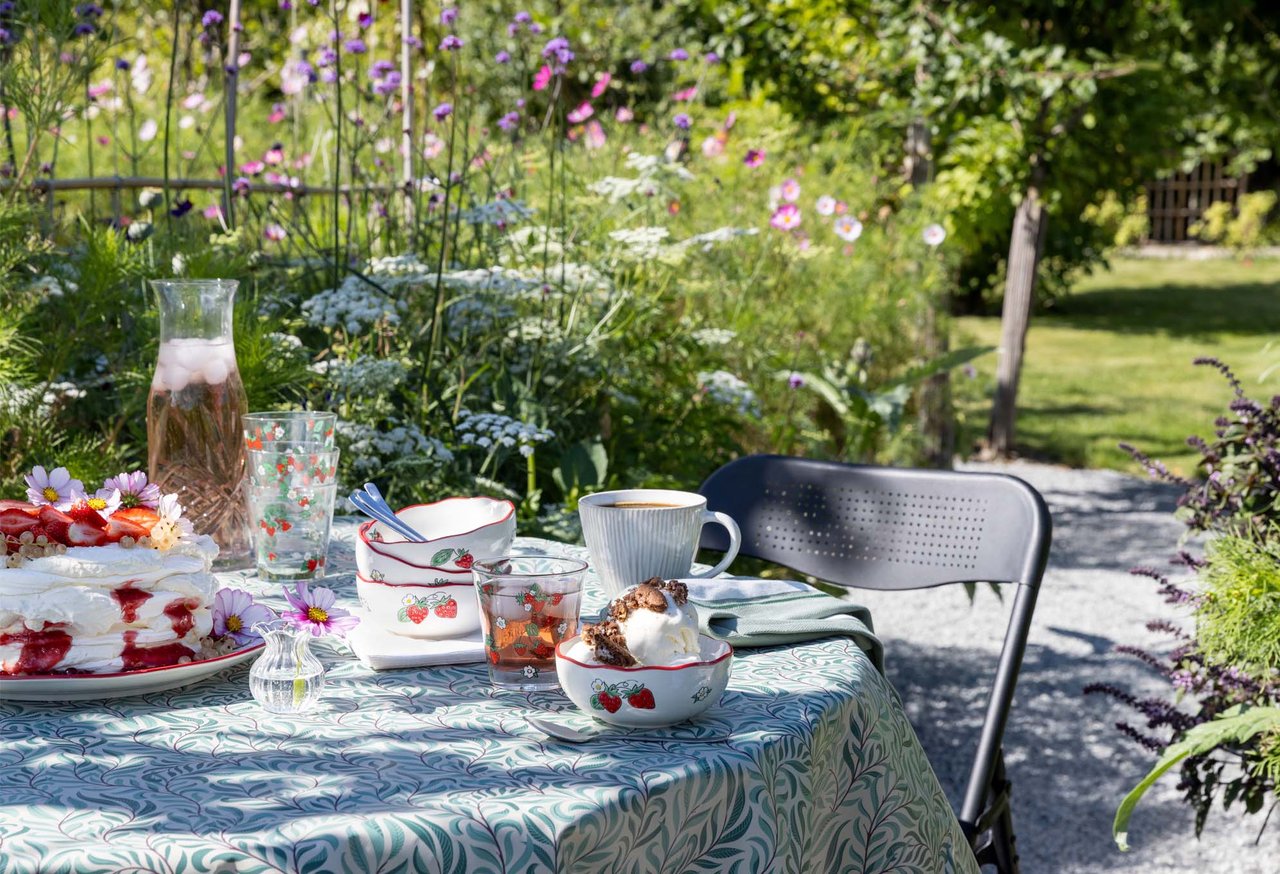 Somrig fikastund i blomstrande trädgård med jordgubbstårta, kaffe och dryck på dukat bord.