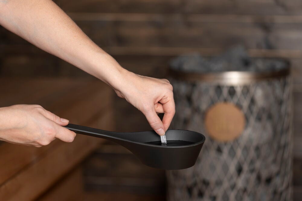 Käsi lisää saunatuoksua mustaan kauhaan lähellä kiuas.