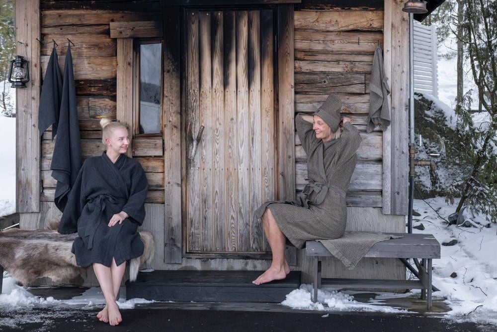 Kaksi henkilöä kylpytakissa saunarakennuksen ulkopuolella laudeliinan kanssa.