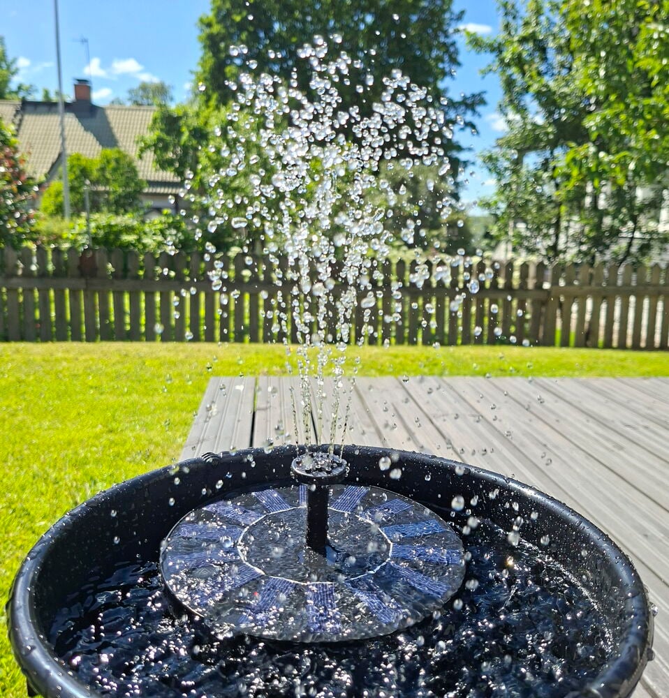 Suihkulähde aurinkokennolla Tarmo, musta pyöreällä sinisellä paneelilla, suihkuttaa vettä ruukussa terassilla.