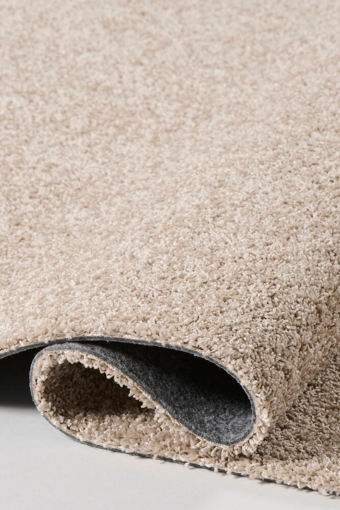 Nærbilde av et beige shaggy polypropen teppe, delvis rullet, som viser den mørkegrå filtbaksiden.