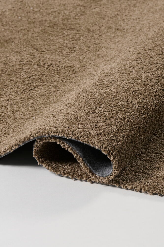 Nærbilde av brunt Teppe Lizette i polypropen, delvis rullet for å vise tekstur og grå bakside.