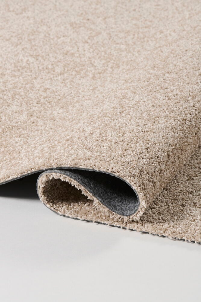 Beige Matto Lizette polypropeenista, osittain rullattu, näyttää sen pörröisen nukan ja tummanharmaan alapuolen.