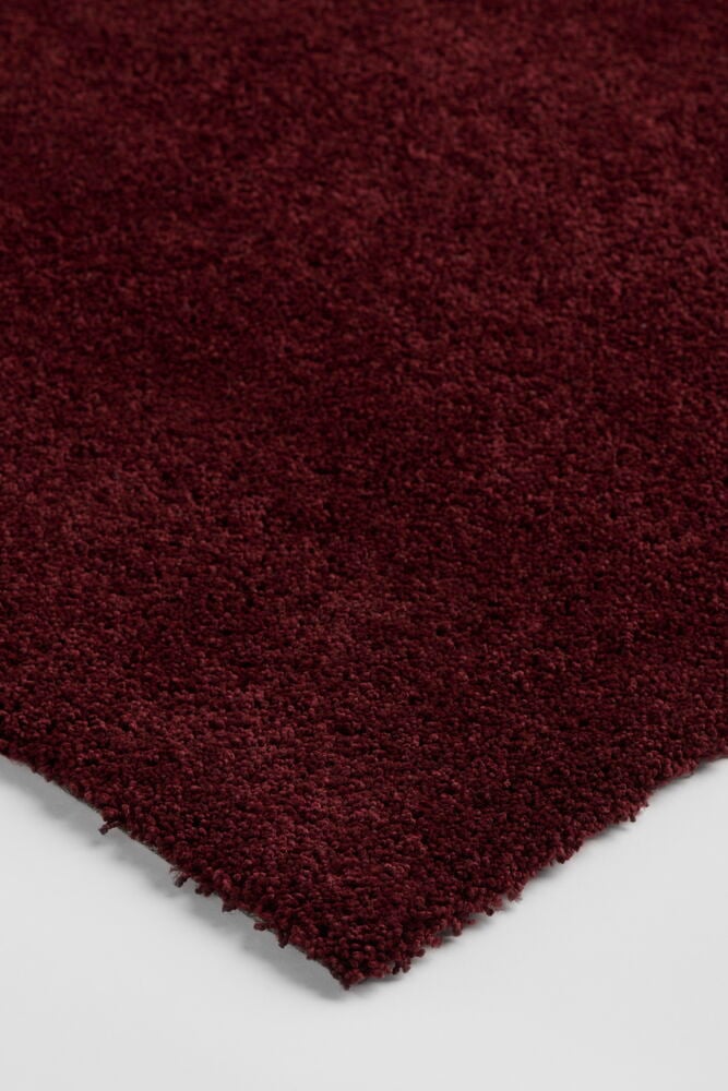 Närbild snett ovanifrån på en lurvig Vinröd Polypropen Matta Lizette med synlig kant på vit yta.