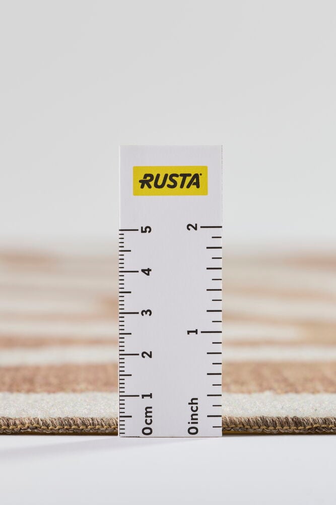 Lähikuva raidallisesta beigen ja kermanvärisestä matosta, jonka paksuutta mitataan valkoisella viivaimella.