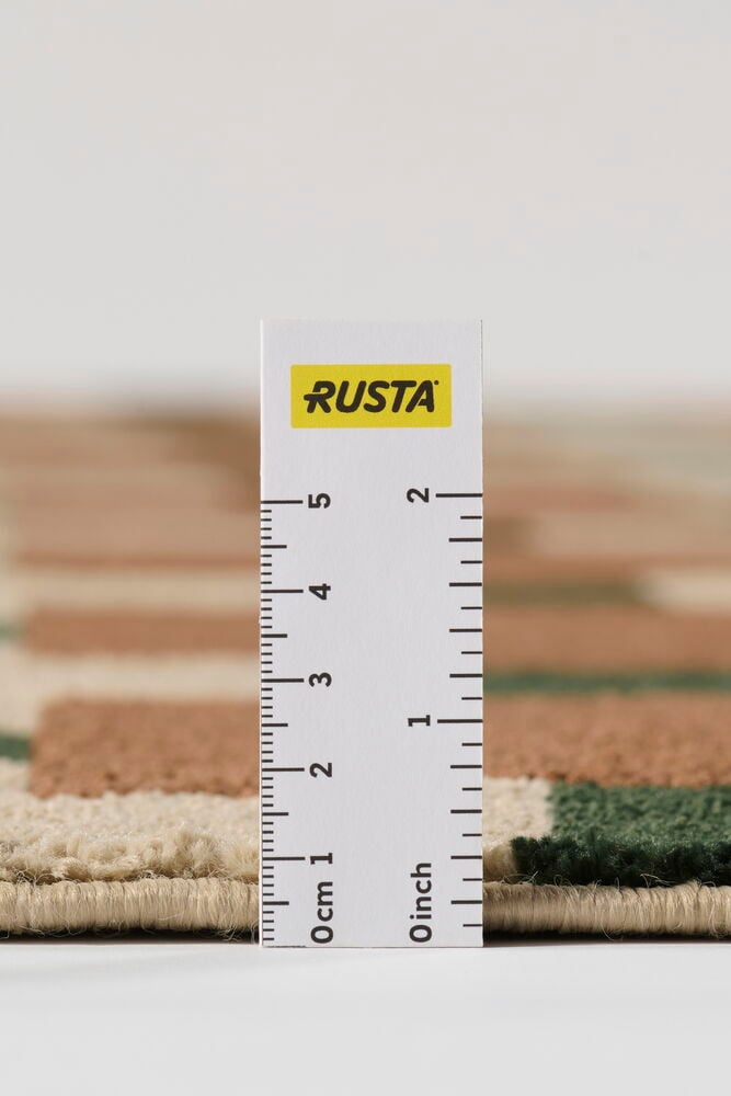 Nærbilde av Teppe Susanna Rust Polyester med beige, brunt og grønt blokkmønster, tykkelsen er målt med en linjal.