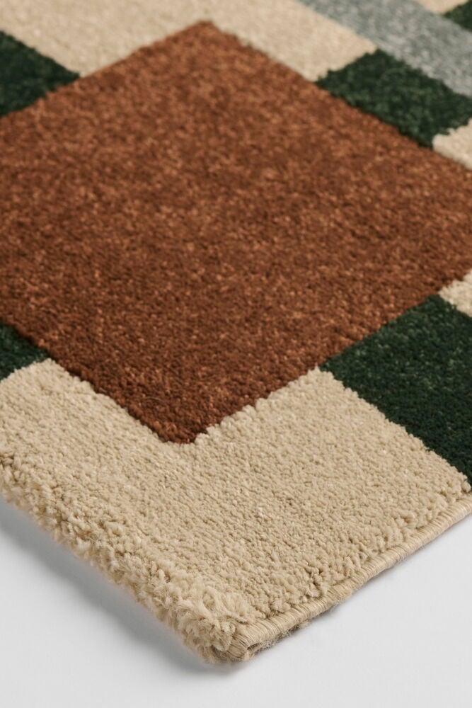 Nærbilde av Teppe Susanna, et polyesterteppe med rust, mørkegrønne og beige geometriske blokker.