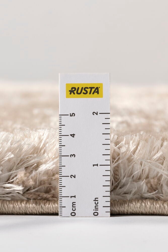 Lähikuva beigen nukkamaton nukan korkeudesta mitattuna valkoisella viivaimella, joka näyttää senttimetrit ja tuumat.