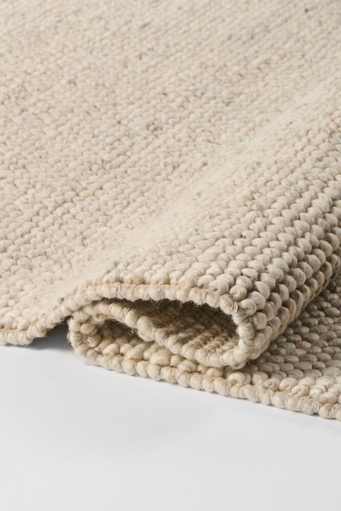 Närbild på en naturfärgad beige ullblandningsmatta med grov, texturerad väv, delvis vikt.