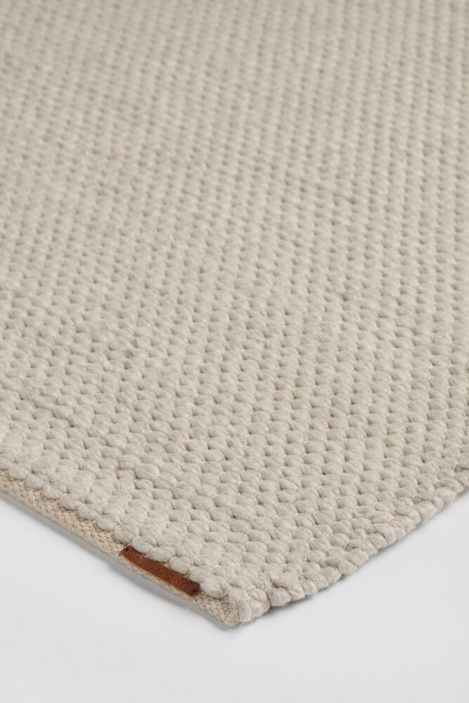 En ljus naturfärgad matta i ullblandning med vävd textur och en liten brun läderetikett.