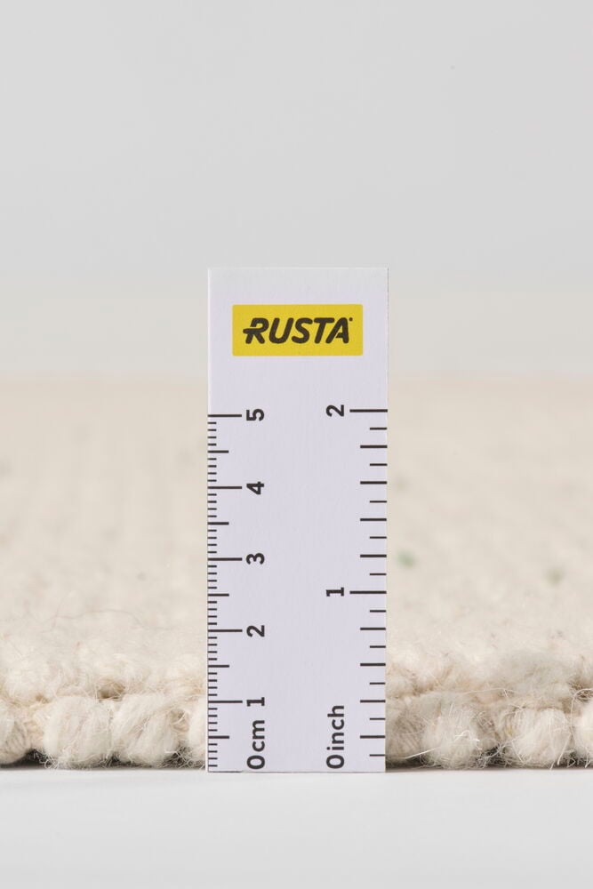 Närbild av en vit ullblandningsmatta som visar textur och flätad kant, med en mätlinjal för skala.