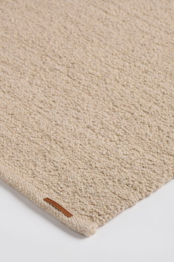 Närbild på beige Elsaform Bonnie matta i ullblandning med texturerad yta och en brun lädertagg.