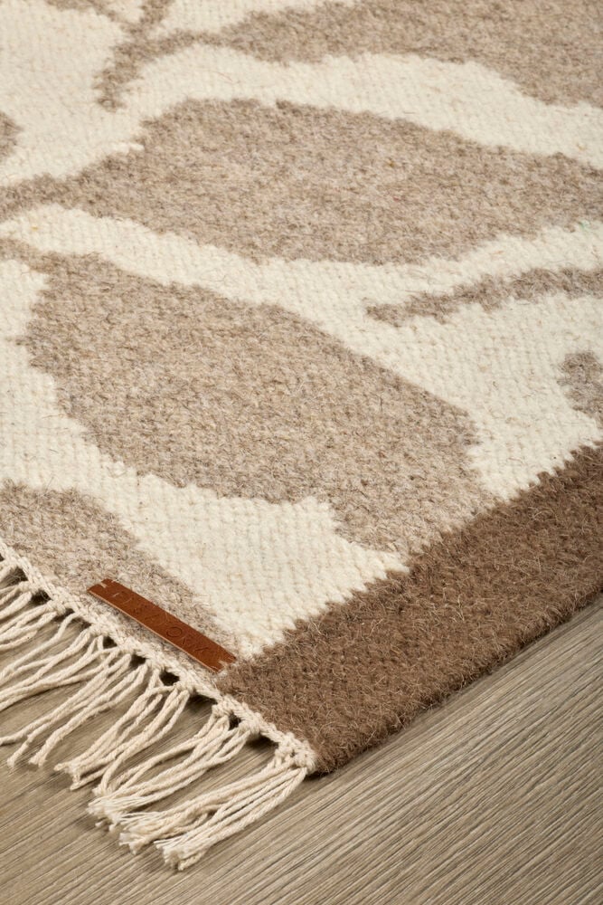 Närbild av en Beige Ull-matta med abstrakt mönster, ljusa fransar och en liten brun etikett.