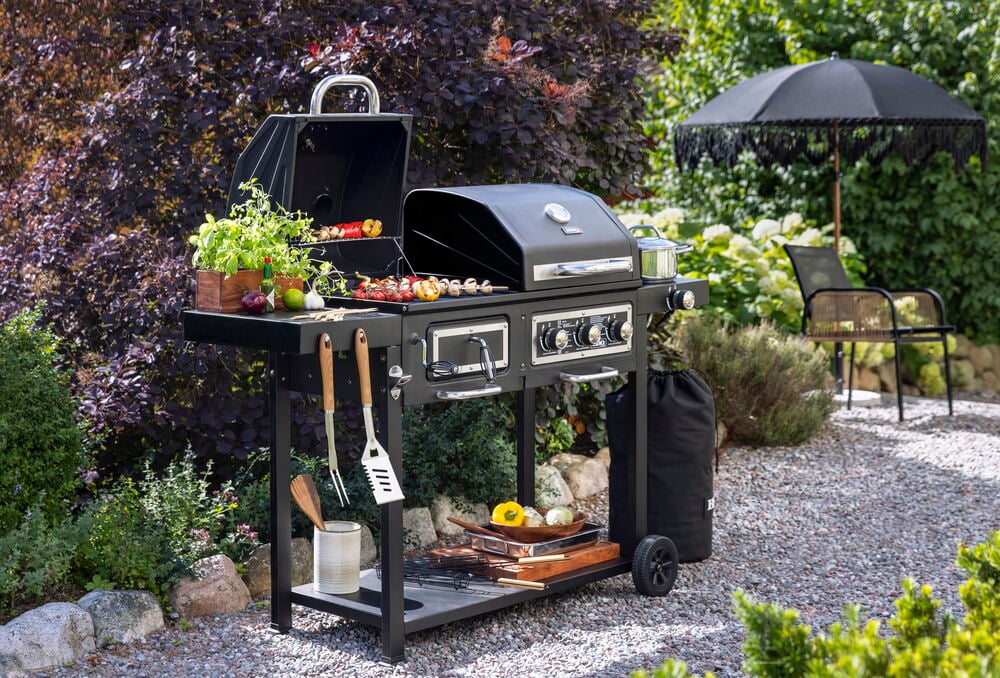 Schwarze Grillplatte, Propangas Sarek mit zubereiteten Speisen, hängenden Utensilien und Kochtöpfen im Garten.