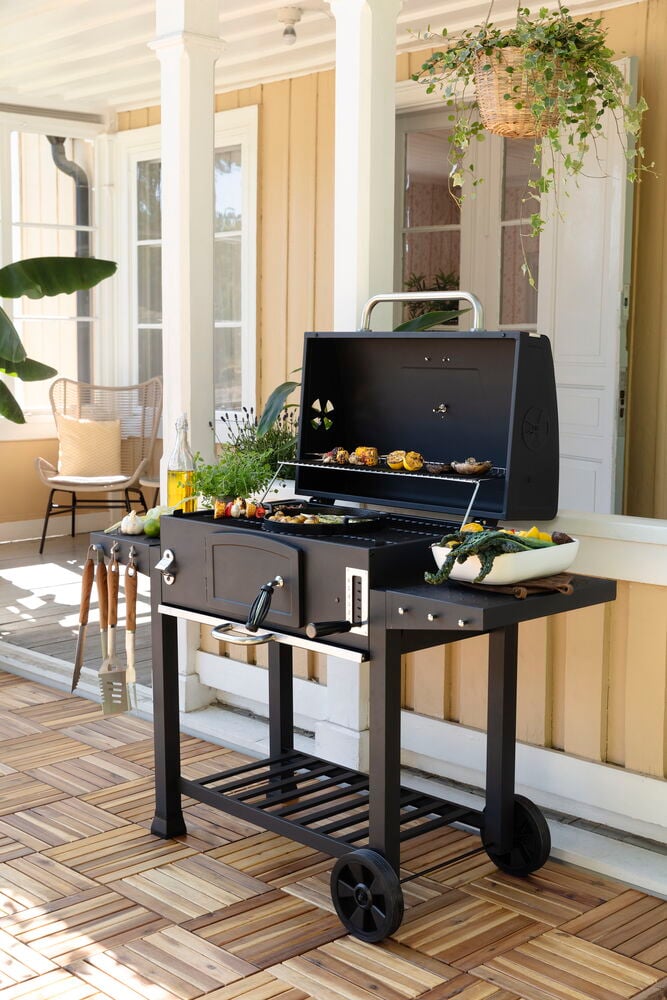 Svart Kullgrill Pampas XL med mat på rister, sidehyller og grillredskaper på en platting.