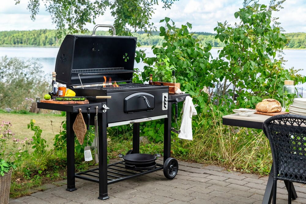 Kullgrill Pampas XL 142x61x116 cm Svart med åpent lokk og flammer, på en uteplass ved innsjø.