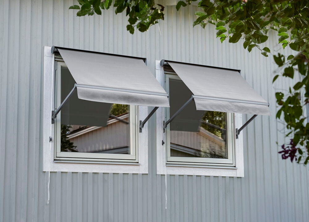 Zwei graue Fenstermarkisen sind auf einem hellgrauen, vertikal getäfelten Haus ausgefahren.