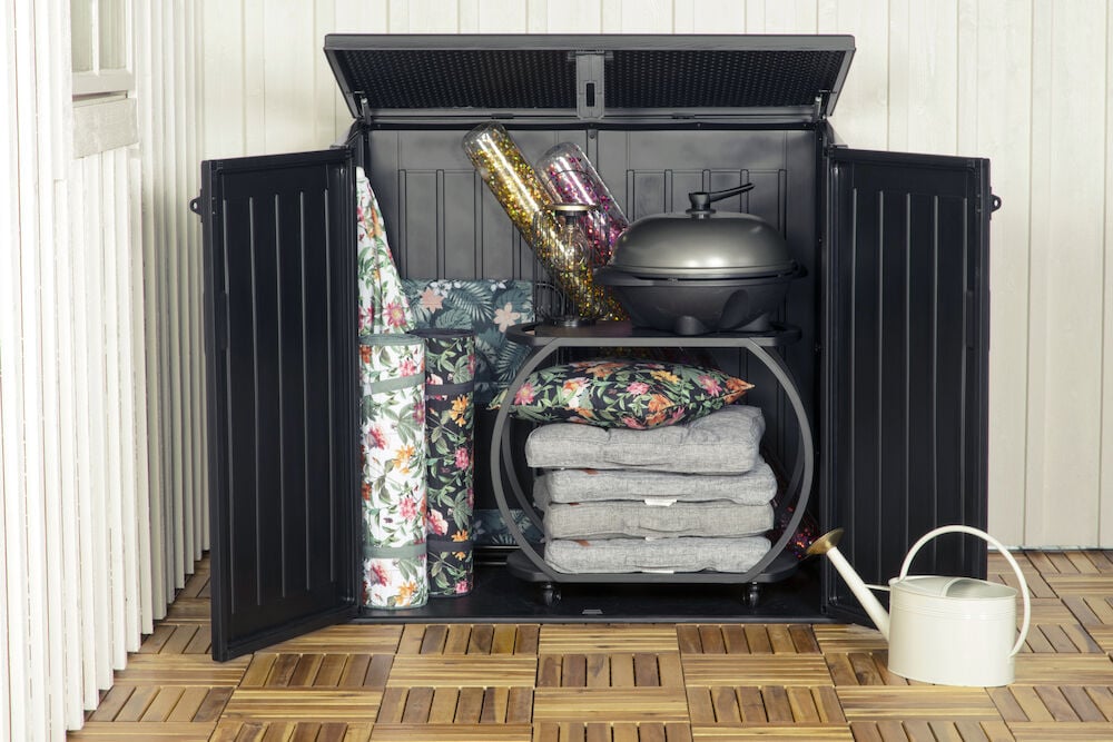 Öppet trädgårdsskjul med grill, dynor och vattna kanna.