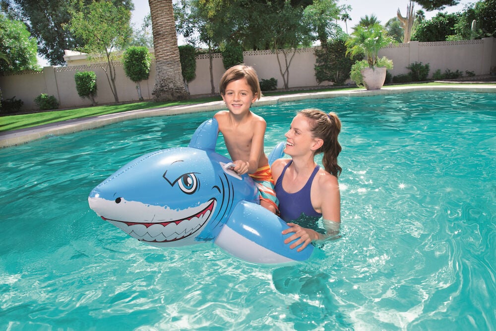 Kind reitet blau-weißes Aufblasbares Wasserspielzeug Shark 157x71 cm, gehalten von Frau im marineblauen Badeanzug.