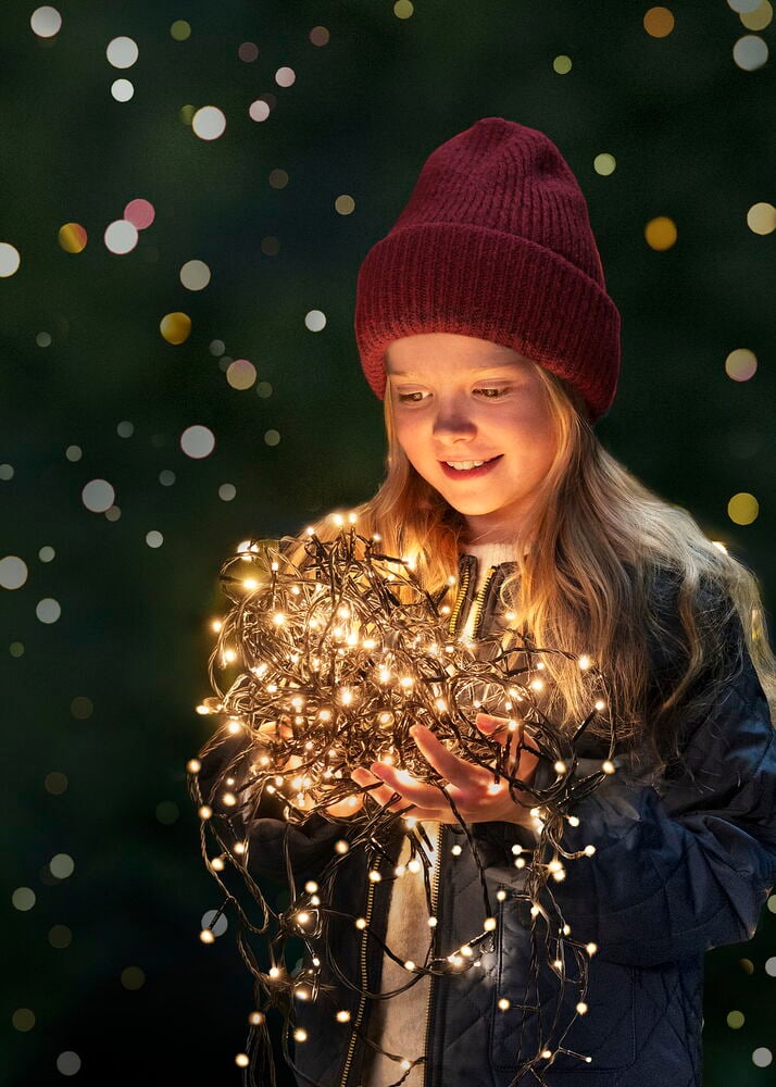 Ett barn håller en Glänsa varmvit 16 LED ljusslinga, med röd stickad mössa och mörk quiltad jacka.