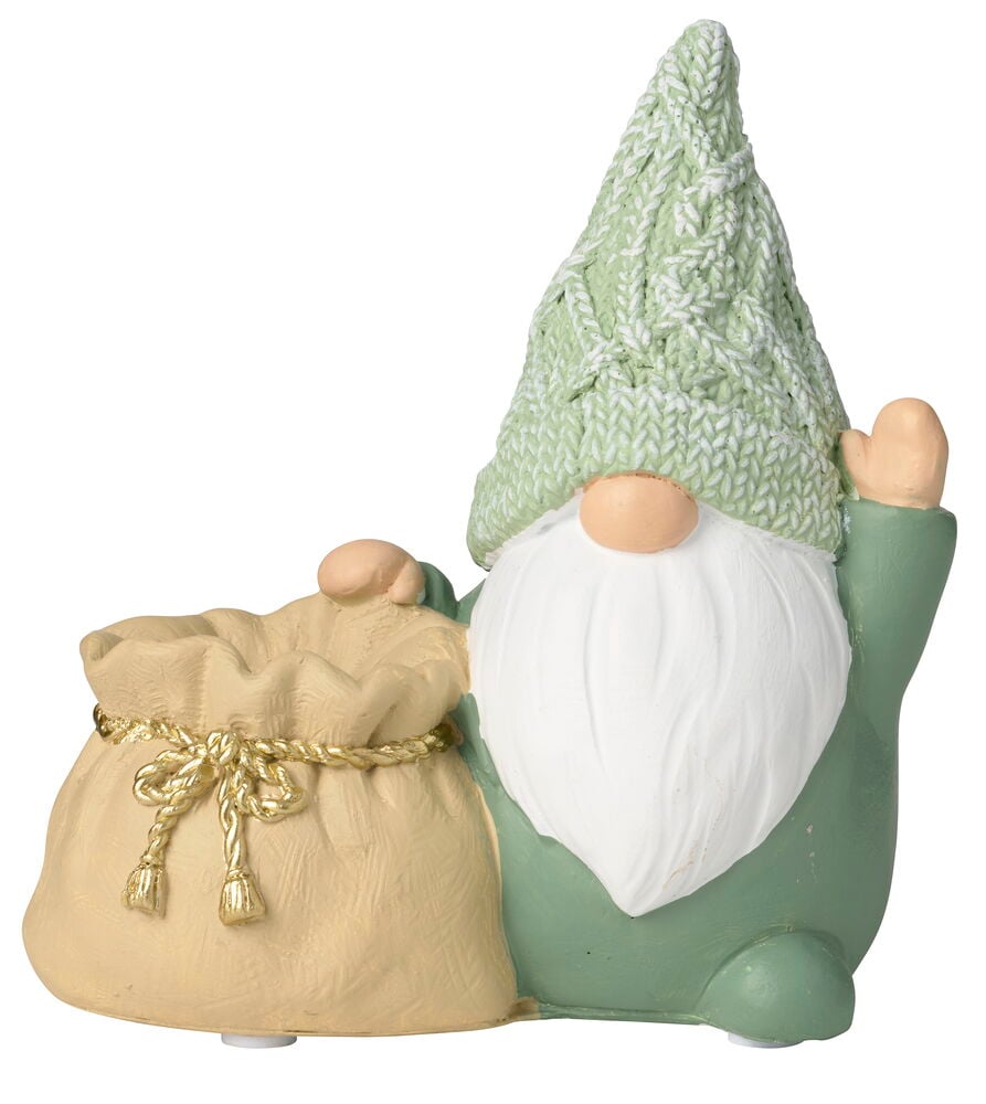 En grön tomte-julljusstake med stickad mössa och vitt skägg, håller en beige säck med guldsnöre.