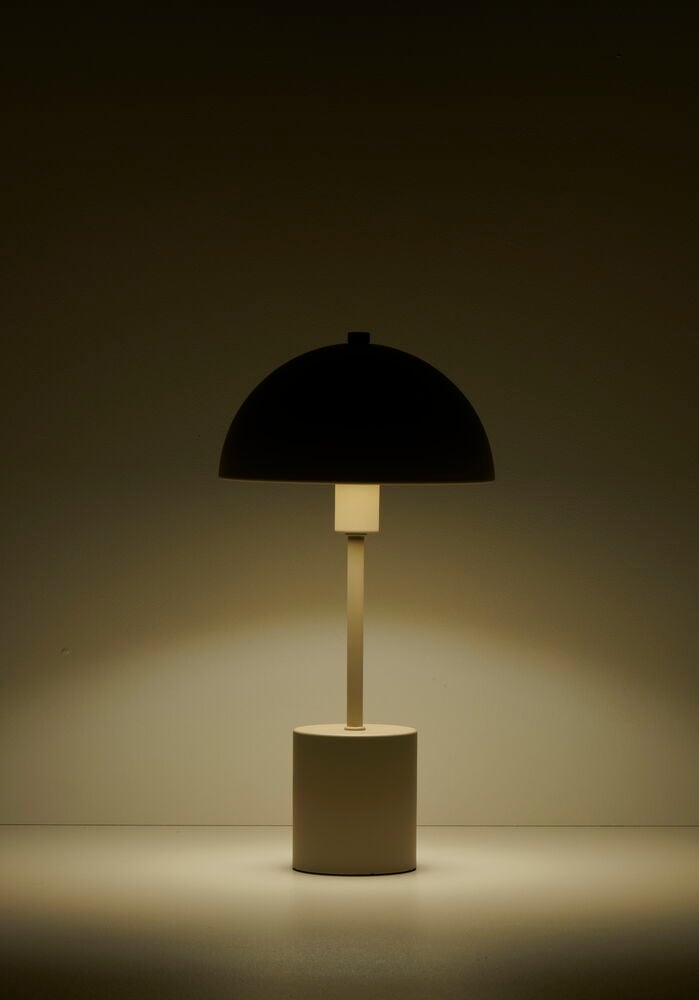 Beige pöytälamppu, jossa pyöreä varjostin, sytytetään tummaa seinää vasten.