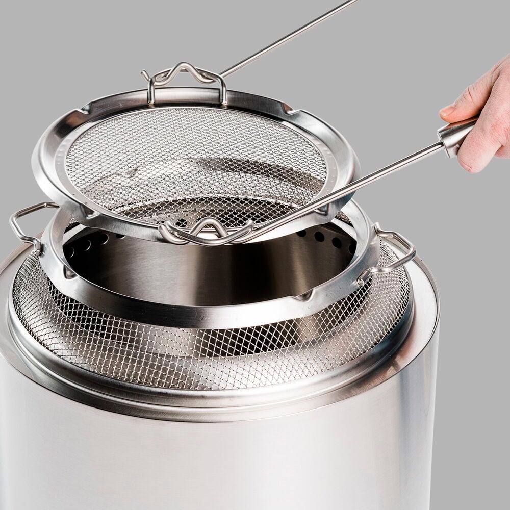 Hand håller avtagbart galler över silverfärgad eldstad.