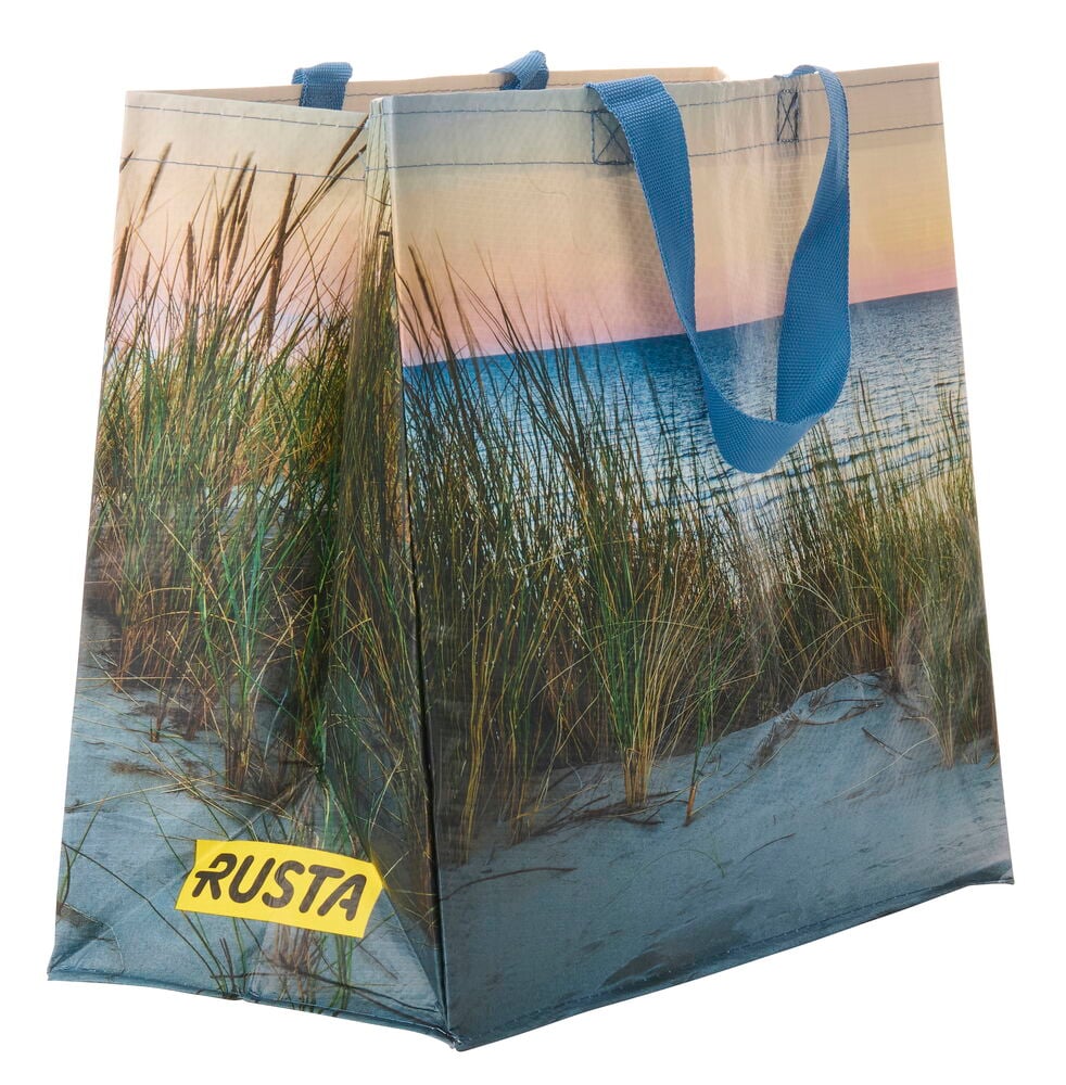 Ostoskassi Medium Kesä, jossa on hiekkadyynien, meren ja auringonlaskun printti sekä siniset kahvat.