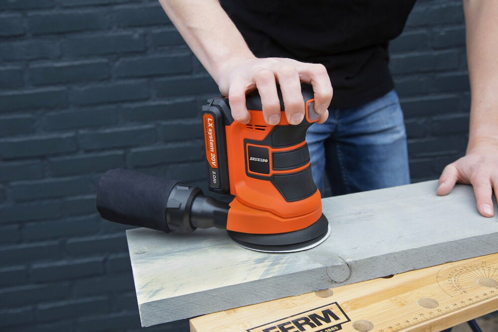 Person slipar träskiva med orange batteridriven slipmaskin.