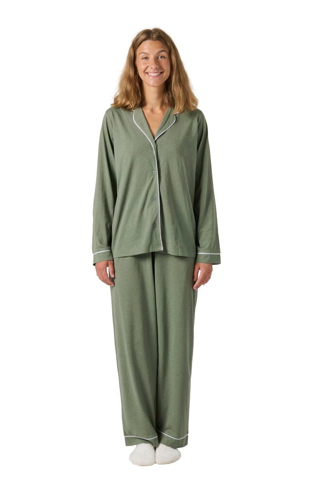 Kvinna bär gröna Pyjamas Laura med vit passpoal, långärmad topp och byxor, med vita tofflor, framifrån.