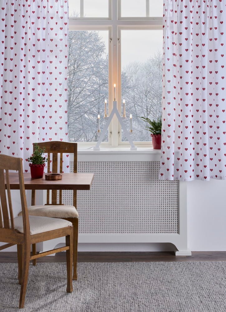 En 2-pakning hvite Gardinlengder Hearts 110x160 cm i bomull med røde hjerter henger i et vindu.