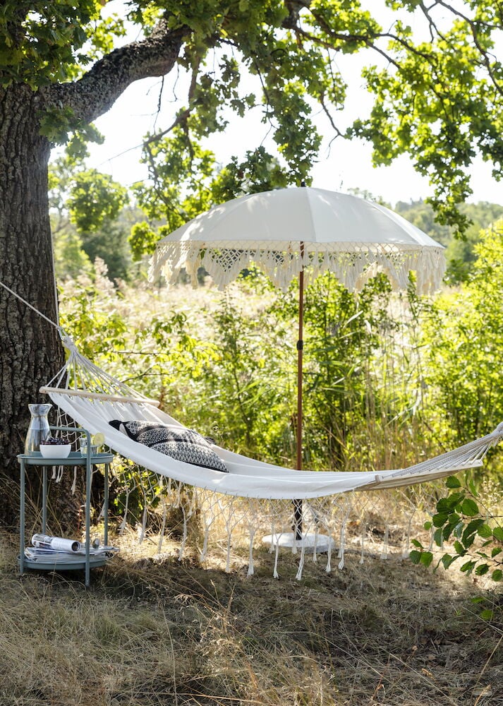 Vit parasoll med tofsar bredvid hängmatta utomhus.