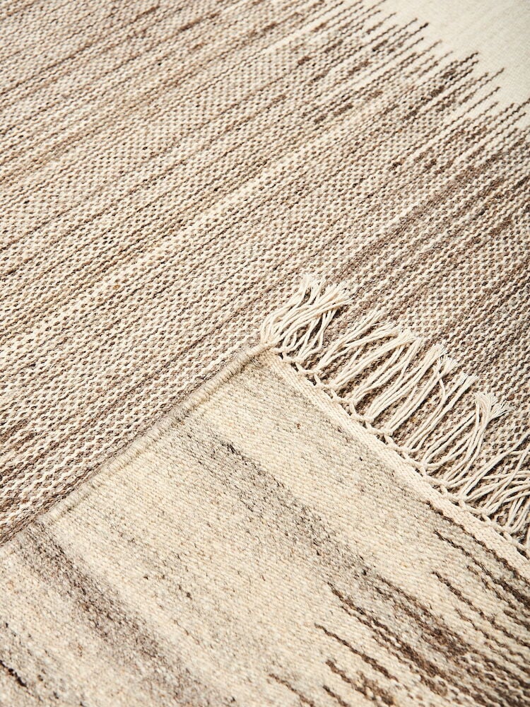 Beige käsinkudottu villamatto, jossa on hapsut ja kirjailtu kuvio.