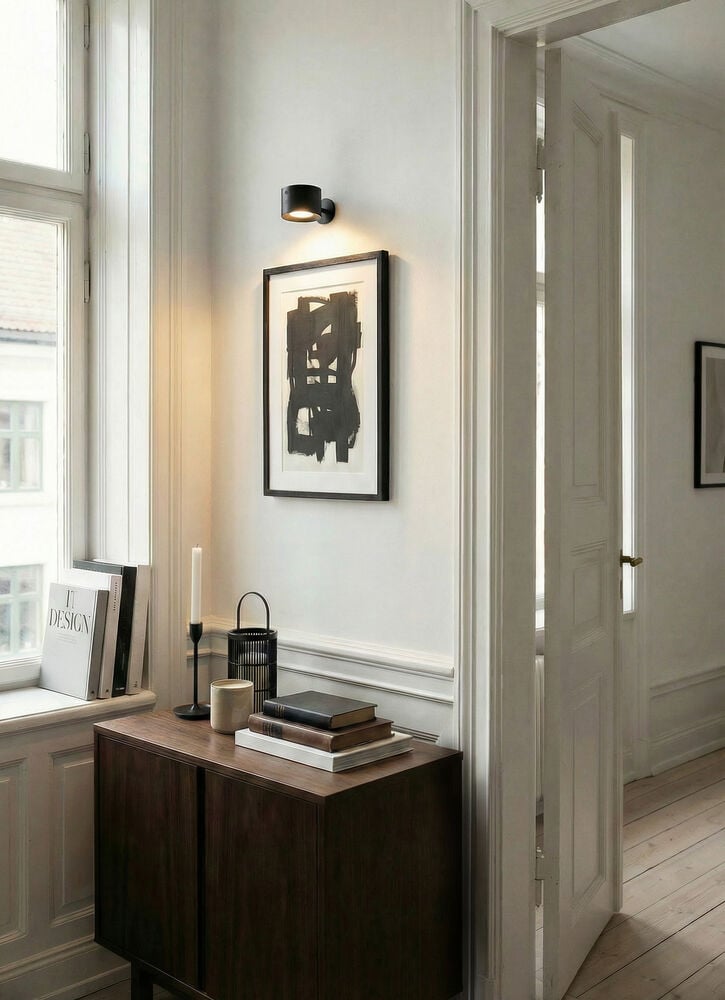 Musta ladattava seinävalaisin valkoisella seinällä abstraktin taiteen ja tumman puukaapin yläpuolella.