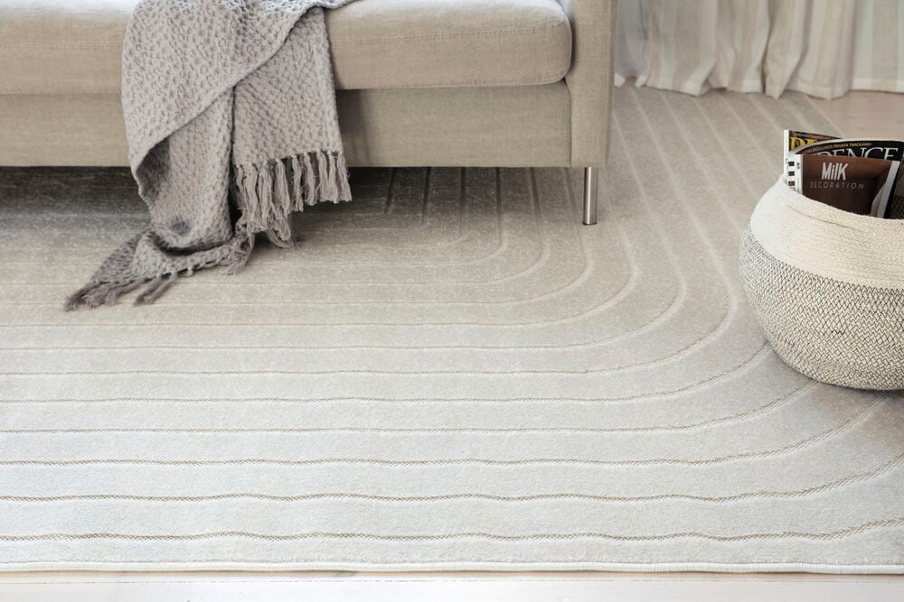 Teppe Billie off-white med bølgede linjer, presentert med beige sofa, grått pledd og flettet kurv.
