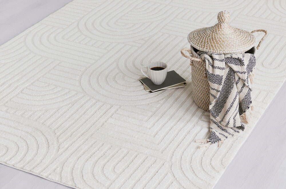 Matta Mette Creme med geometriskt mönster, en flätad korg, svartvit pläd och en kaffekopp.
