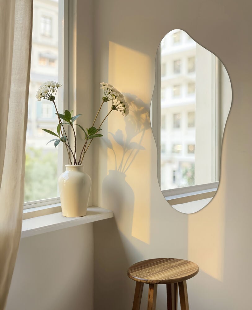 Organiskt formad spegel på ljus vägg vid fönster.