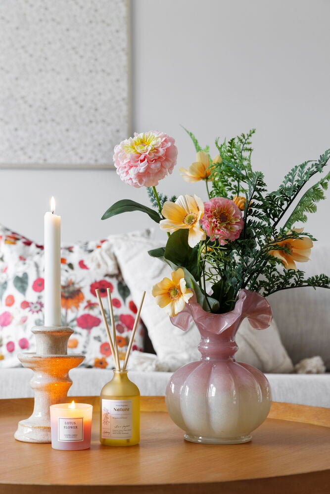 Konstgjord blombukett i rosa vas, med två ljus och en gul doftspridare.