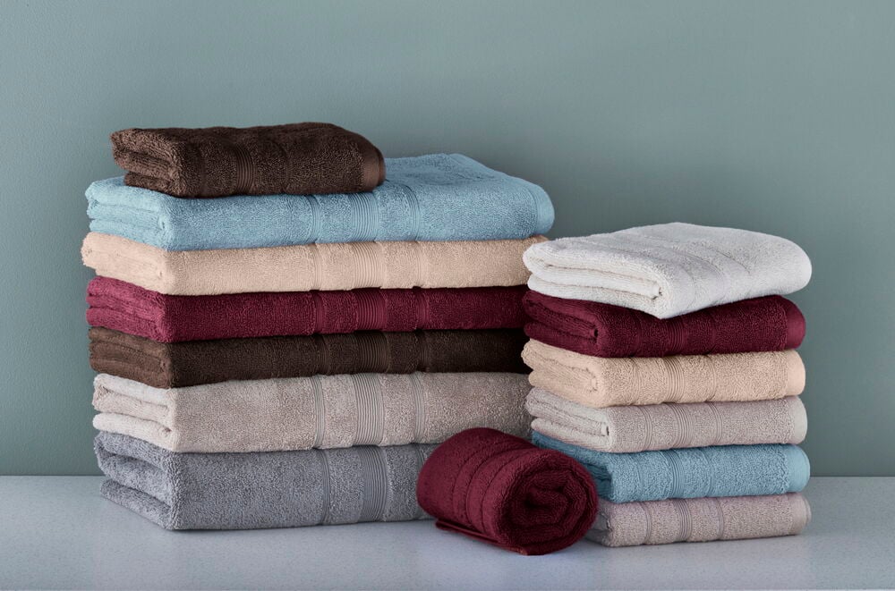 Stapel gefalteter Handtücher in Weiß, Beige, Hellblau, Bordeaux, Braun und Grau, sowie ein gerolltes bordeauxfarbenes Handtuch.