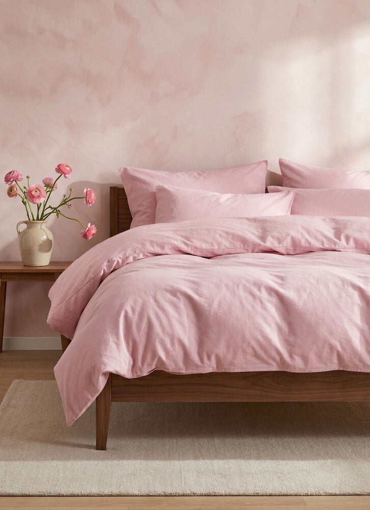 Ein Bett präsentiert ein altrosa Baumwoll Bettwäsche-Set Vida, mit Bettbezug und mehreren Kissenbezügen, auf einem Holzrahmen.