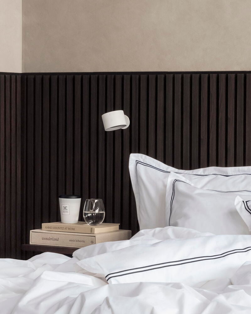 Ladattava seinävalaisin Valkoinen yöpöydän esineiden yllä: lasi, kuppi, kirjat ja valkoiset vuodevaatteet.